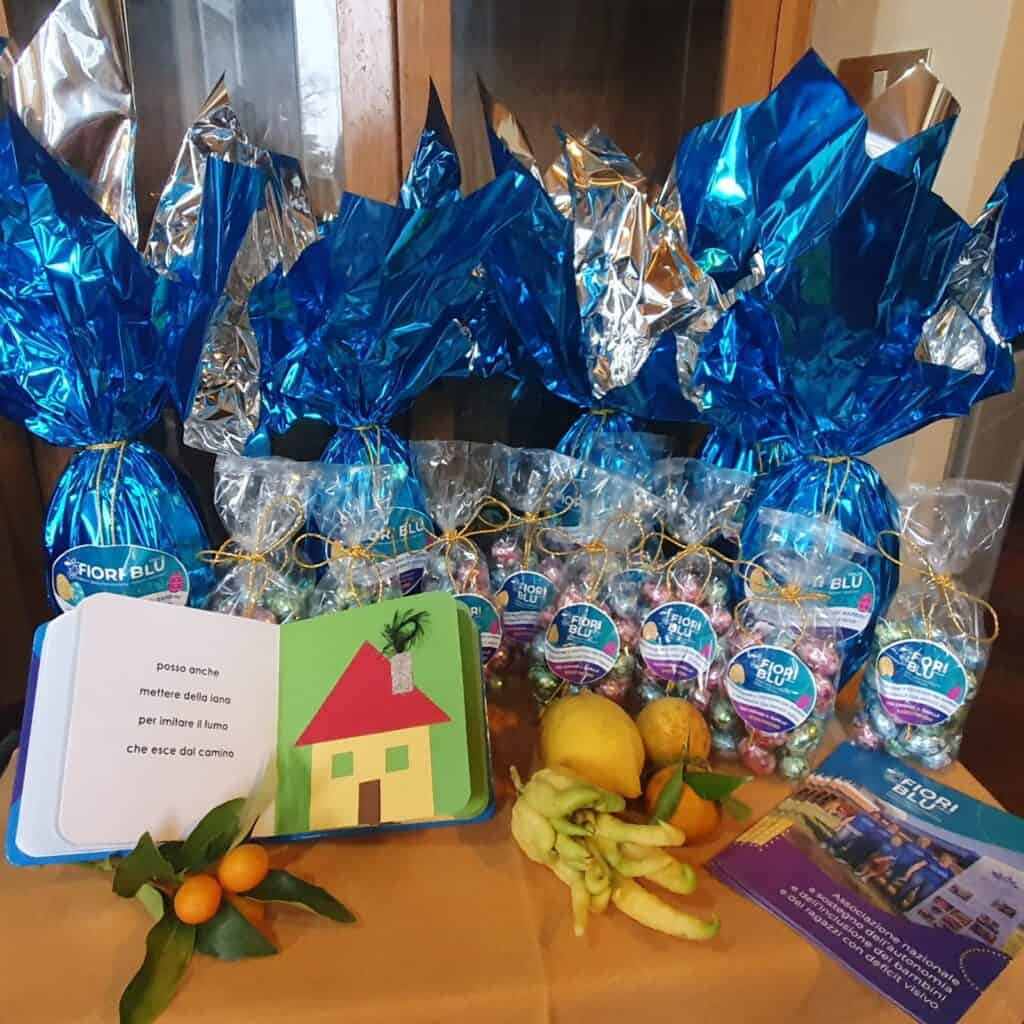 uova e sacchetti di cioccolato dell'associazione fiori blu su un tavolo con depliant, un librino tattile e alcuni agrumi di Cannero