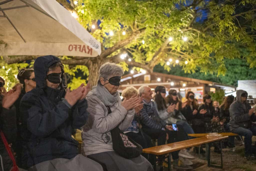 In a park in the evening, some blindfolded spectators sit watching a concert while clapping their hands