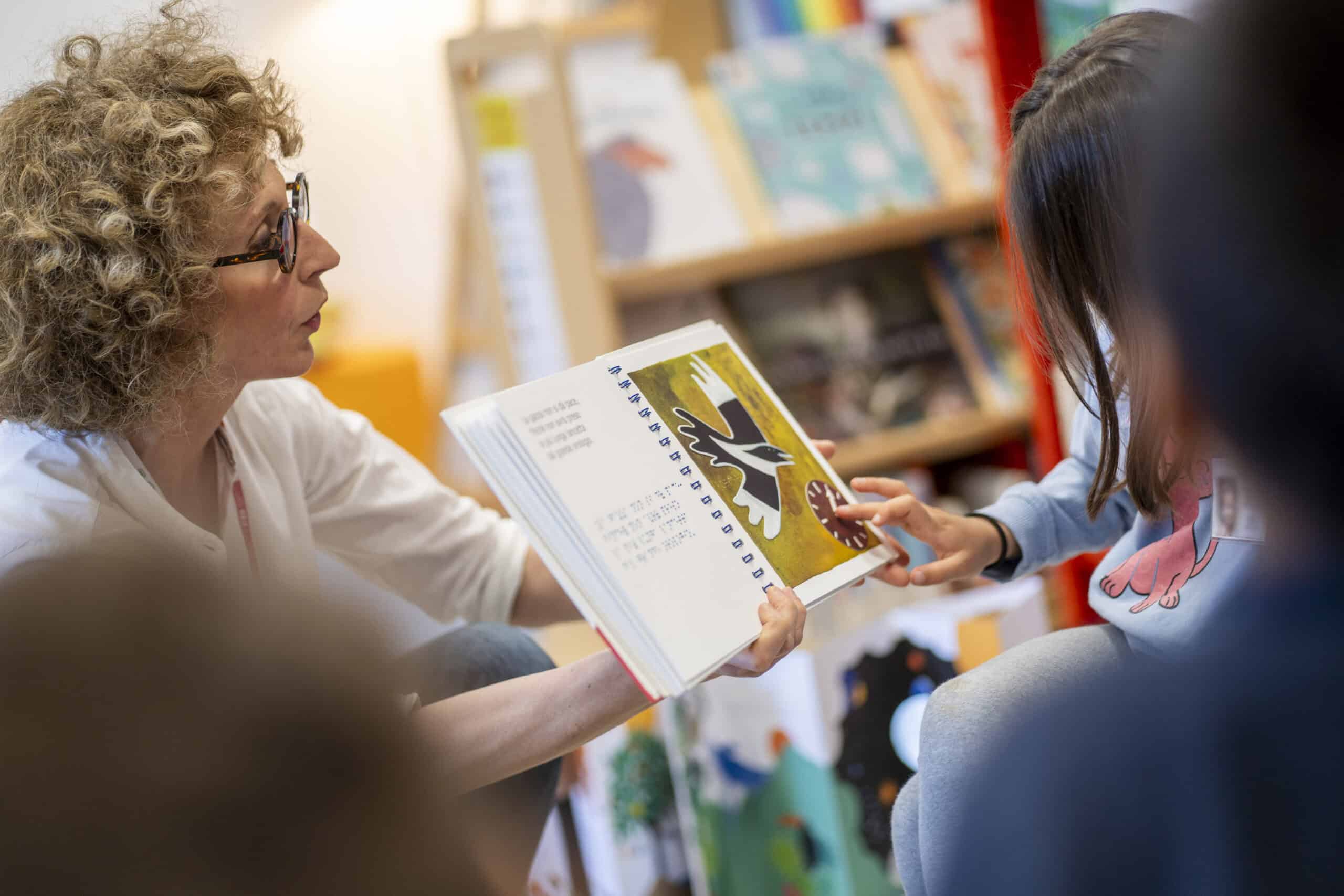Una bibliotecaria apre un libro tattile davanti a dei bambini. Uno di loro tocca con le dita l'immagine di un orologio con le lancette