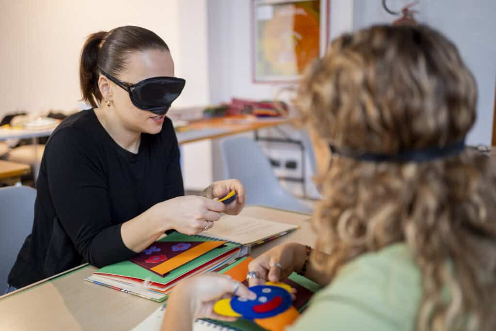 Two blindfolded women touch some fabric figures and silhouettes