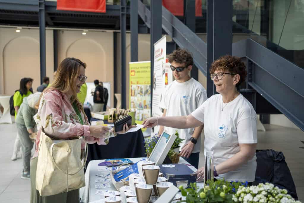 two volunteers behind a banquet explain the activities of the Blue Flowers association to a lady inside the San Gaetano Center in Padua