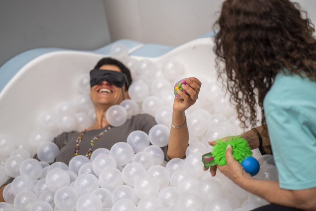 a blindfolded woman is immersed in a tub of transparent balls, and a caregiver hands her some sensory materials