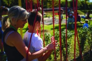 un'educatrice introduce un bambino non vedente all'interno della pista da corsa sensoriale, tra fili di tessuto rosso e campanellini