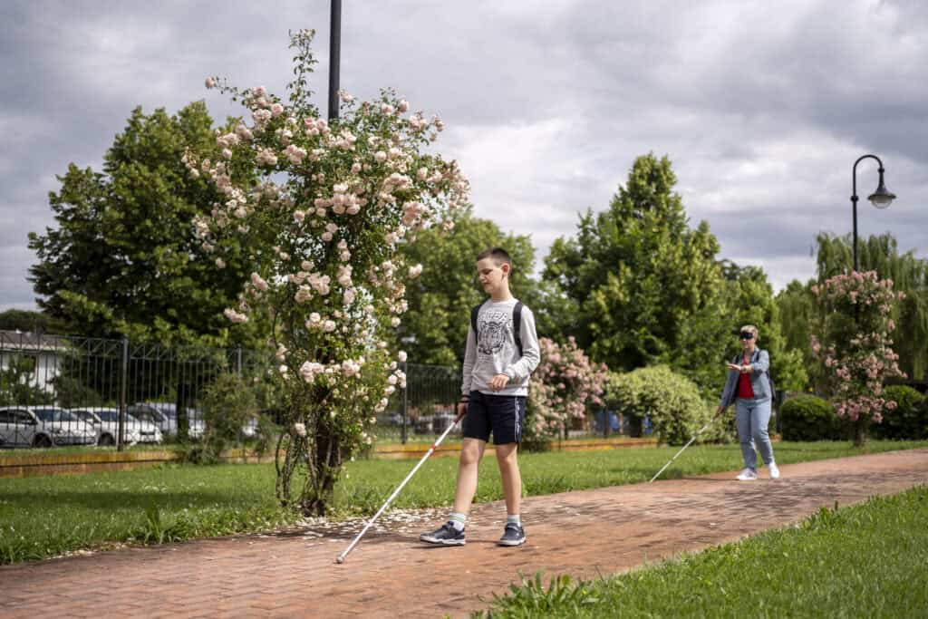 un ragazzo con il bastone bianco cammina lungo il viale del Centro di Padova. Alle sue spalle una donna bendata e con bastone bianco fa esperienza di deprivazione visiva